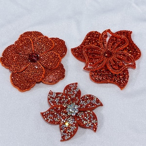 May include: Three flower-shaped appliqués covered in sparkling crystals. Two of the flowers are a deep orange-red color, while the third flower is a vibrant red with a center of clear, reflective crystals that create a bright, shimmering effect.