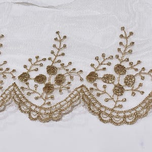 May include: Gold embroidered lace trim with a scalloped edge. The trim features a repeating floral and branch design. The lace is on a white mesh background. This trim is suitable for sewing and craft projects.