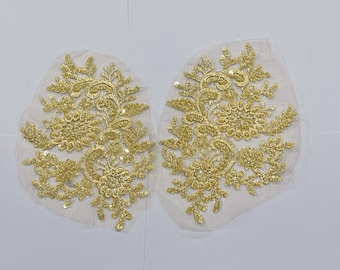May include: Two gold floral lace appliques with sequins on a white mesh background. The appliques are shaped like a flower with a stem and leaves.