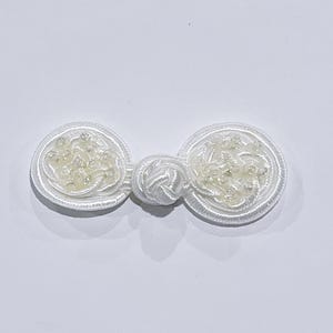 May include: White fabric button with a knotted center and pearl-like embellishments.