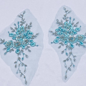 May include: Two matching floral appliques. Each applique features a central cluster of turquoise blue flowers with silver-colored bead and sequin accents. Delicate silver-colored beaded vines and leaves extend from the floral clusters, all on a sheer, light blue fabric.
