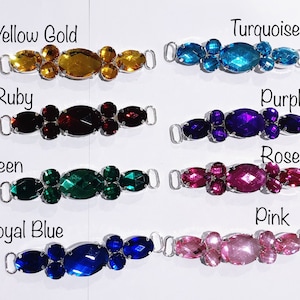 May include: A collection of colorful rhinestone embellishments in various colors, including yellow gold, ruby, green, royal blue, turquoise, purple, rose, and pink. Each piece has a silver-tone metal loop on each end and is labeled with its color.