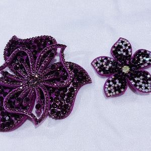 May include: Two flower-shaped appliques in shades of purple. The larger flower features dark purple crystals on a purple base. The smaller five-petaled flower is decorated with a mix of dark purple and clear crystals, with a single clear center stone.