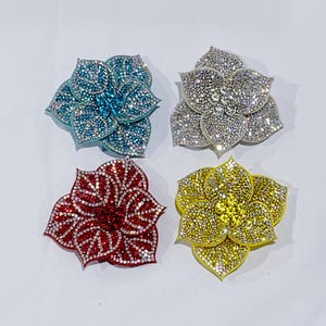 May include: Four flower-shaped brooches with sparkling rhinestone embellishments. The brooches are in shades of blue, silver, red, and yellow. Each brooch has multiple petals and a central cluster of rhinestones.