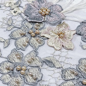 May include: Close-up of delicate floral lace with embroidered silver and pale pink flowers. Each flower is adorned with small, round, pearl-like beads. White feathers accent some of the flowers, creating a soft, elegant texture. The lace is on a white background.