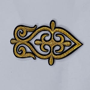 May include: A gold-colored embroidered patch with a black border. The patch features a symmetrical, ornate design with swirling patterns and a pointed end, suitable for embellishing clothing or accessories.