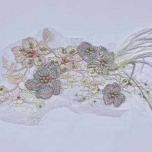 May include: A delicate, floral-themed applique featuring embroidered flowers in shades of pink, gold, and silver, adorned with pearl-like beads. White feathers extend from the design, creating an elegant, decorative piece.
