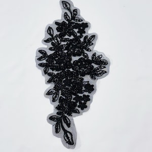 May include: Black floral applique with beaded and sequined details. The applique features a cluster of flowers and leaves, all in a dark, solid color. The design is set on a sheer, gray backing.