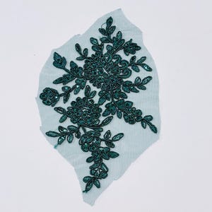 May include: A teal-colored fabric applique with a floral design. The applique features dark teal embroidered leaves and flower-like shapes, embellished with sequins. The applique is on a light blue mesh backing.