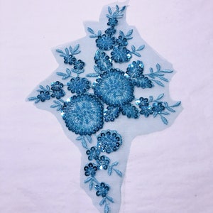 May include: A light blue floral applique with sequins and beads on a sheer mesh fabric. The applique features a large flower with smaller flowers and leaves surrounding it.