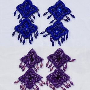 May include: Four pairs of beaded earrings in shades of blue and purple. Each earring features a diamond-shaped design with a central jewel and beaded fringe. The earrings are likely handmade and suitable for special occasions.
