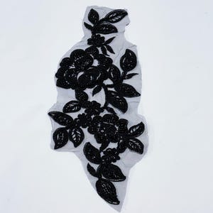 May include: Black floral applique on a gray mesh backing. The applique features detailed black flowers and leaves, with a delicate, textured appearance. The design is intricate and appears to be hand-stitched, suitable for embellishing garments or crafts.