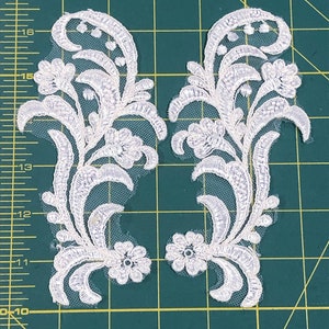 May include: Two white lace appliques with floral designs. The appliques are about 12 inches tall and have a delicate, intricate pattern.