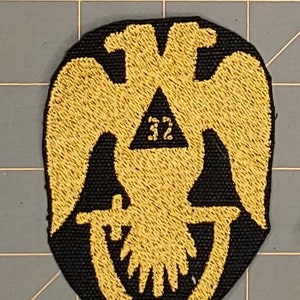 May include: A black and gold embroidered patch featuring a double-headed eagle with a triangle containing the number "32" in the center. The eagle is holding a crescent moon in its talons.