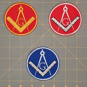 Könnte beinhalten: Drei gestickte Aufnäher mit dem freimaurerischen Symbol eines Zirkels und eines Geodreiecks mit einem 'G' in der Mitte. Die Aufnäher sind rot, gelb und blau.