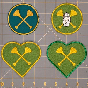 Può includere: Quattro toppe ricamate in verde e giallo. Due toppe sono a forma di cuore e due sono circolari. Le toppe presentano un disegno di due remi incrociati con una mano al centro di una delle toppe circolari.