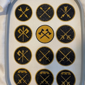 Puede incluir: Una tela blanca con 12 círculos bordados en negro y dorado. Cada círculo contiene un diseño diferente de armas cruzadas, incluyendo espadas, hachas, lanzas y flechas.