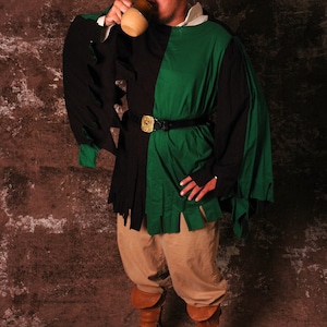 May include: A man wearing a black and green tunic with a brown belt and brown boots. He is holding a brown ceramic mug and drinking from it.