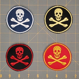 May include: Four embroidered iron-on patches featuring a skull and crossbones design. The patches are circular and come in four color combinations: black with white skull and crossbones, black with gold skull and crossbones, red with gold skull and crossbones, and blue with white skull and crossbones.