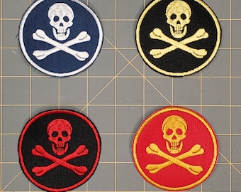 May include: Four embroidered iron-on patches featuring a skull and crossbones design. The patches are circular and come in four color combinations: black with white skull and crossbones, black with gold skull and crossbones, red with gold skull and crossbones, and blue with white skull and crossbones.