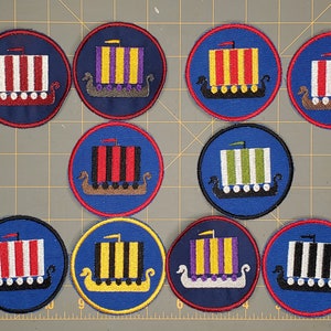May include: Nine embroidered iron-on patches featuring a Viking ship with different colored stripes. The patches are circular and have a red outer border.