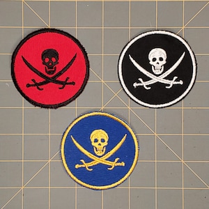 Op de afbeelding: Drie ronde patches met een doodshoofd en gekruiste botten. Eén patch heeft een rode achtergrond met een zwarte schedel en gekruiste zwaarden. Een andere heeft een zwarte achtergrond met een witte schedel en zwaarden. De derde heeft een blauwe achtergrond met een gouden schedel en zwaarden.