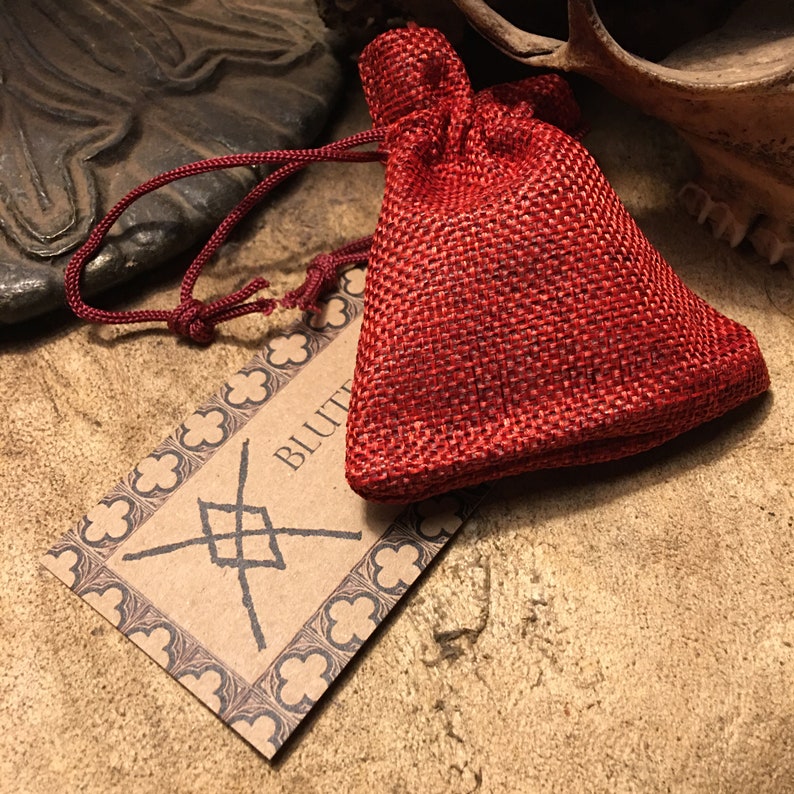 May include: A small red burlap pouch with a drawstring closure. The pouch is sitting on a brown surface with a tag that says "BLUTT" and has a symbol on it.