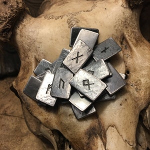 May include: A collection of metal runes, each with a different symbol, arranged on a textured surface. The runes are silver in color and have a worn, aged appearance.