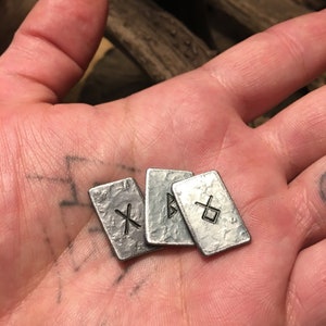 May include: Four silver metal tokens with different symbols etched on them. The tokens are arranged in a pile on a hand.