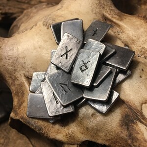 May include: A set of metal runes with a hammered finish. The runes are arranged in a pile on a textured surface. The runes are silver in color and have black markings.