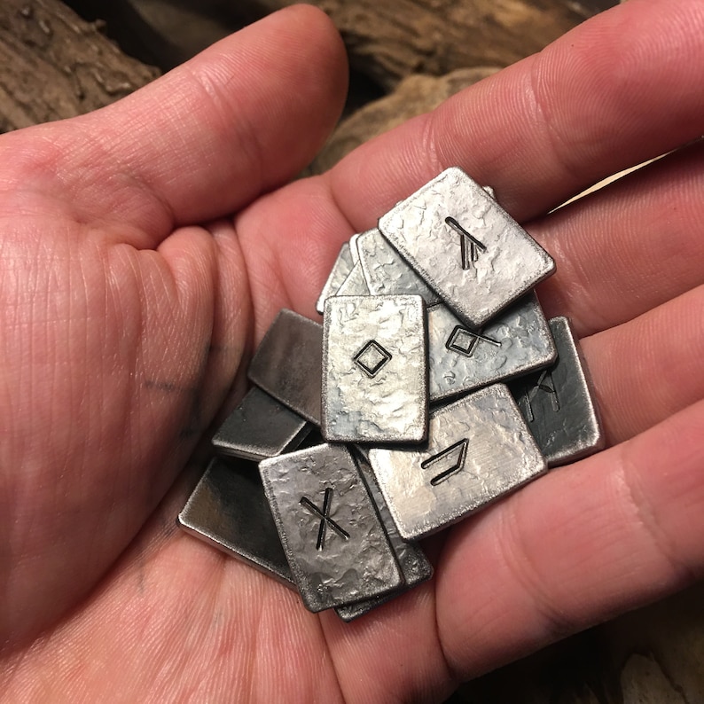 May include: A collection of metal rune tiles with various symbols engraved on them. The tiles are silver in color and have a textured surface.