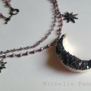 May include: A layered necklace featuring a crescent moon pendant with a dark, textured surface and a silver-colored edge. The necklace also includes star-shaped charms and a beaded chain in shades of purple.