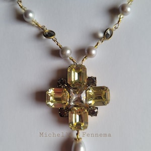 May include: A gold-toned necklace featuring a cross pendant with large, yellow-green rectangular crystals and smaller, dark brown crystals. The necklace includes a dangling pearl and is strung with pearls and small, gray-toned stones.