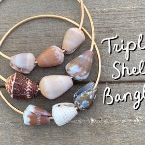 May include: Gold-tone bangle bracelet featuring three strands, each adorned with cone-shaped seashells in shades of brown, cream, and white. The text "Triple Shell Bangle" is written in a script font.
