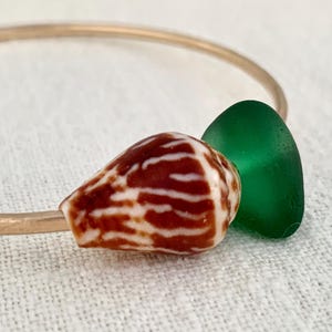 May include: A gold-coloured bangle bracelet featuring a brown and white seashell bead and a green sea glass bead. The bracelet rests on a white, textured surface.