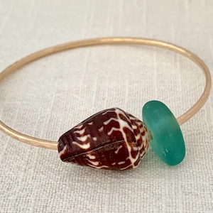 May include: A gold-coloured bangle bracelet with a brown and white seashell and a teal sea glass bead. The bracelet has a hammered texture and a simple design. The bracelet is suitable for everyday wear.