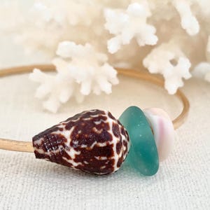 May include: A gold-coloured bangle bracelet featuring a brown and white seashell, a teal sea glass piece, and a pink and white shell. The bracelet is set against a backdrop of white coral.