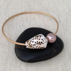 May include: A gold-colored bangle bracelet featuring a white seashell with brown spots and a pink pearl accent. The bracelet is resting on a black stone, set against a neutral background.
