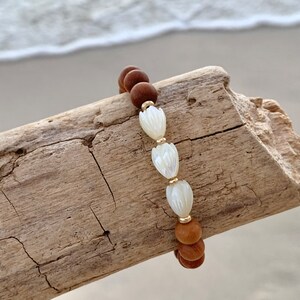 Peut inclure: Un bracelet en bois brun perl&eacute; avec trois perles de coquillage blanches. Le bracelet est sur un morceau de bois flott&eacute;.