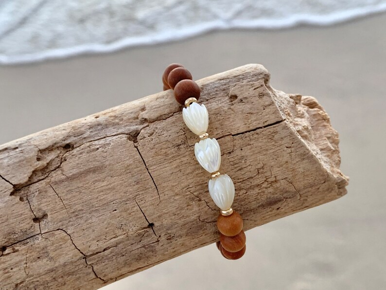 Peut inclure: Un bracelet en bois brun perl&eacute; avec trois perles de coquillage blanches. Le bracelet est sur un morceau de bois flott&eacute;.