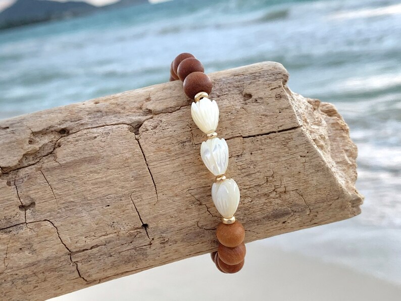 Peut inclure: Un bracelet en bois brun avec trois perles de fleurs en coquillage blanc et des accents dor&eacute;s.