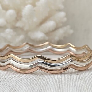 May include: Three wavy rings stacked together. The top ring is gold, the middle ring is silver, and the bottom ring is rose gold.