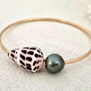 May include: A gold-tone bangle bracelet featuring a seashell with brown spots and a dark gray pearl. The bracelet is a simple, elegant design, perfect for everyday wear. The bracelet's diameter is approximately 2.5 inches.