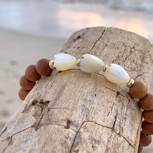 Peut inclure: Un bracelet en bois brun avec des perles de fleurs en coquillage blanc et des accents dor&eacute;s.