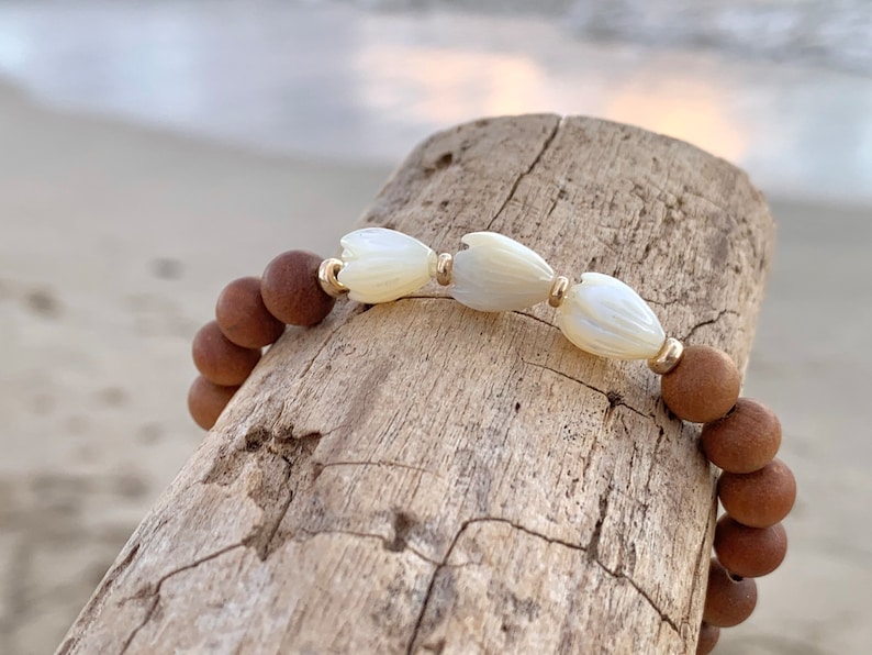 Peut inclure: Un bracelet en bois brun avec des perles de fleurs en coquillage blanc et des accents dor&eacute;s.