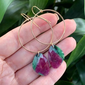 May include: Gold hoop earrings with teardrop-shaped ruby and green gemstone pendants. The earrings are held in a hand, with a green leafy background. The gemstones have a faceted cut, and the gold hoops are thin and delicate.