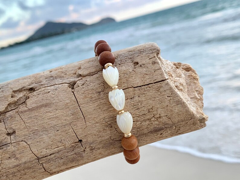 Peut inclure: Un bracelet en bois brun avec des accents de fleurs en coquillage blanc. Le bracelet est sur un morceau de bois flott&eacute;.