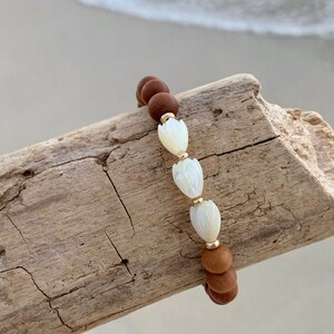 Peut inclure: Un bracelet en bois brun perl&eacute; avec trois perles de fleurs en coquille blanche. Le bracelet est sur un morceau de bois flott&eacute;.