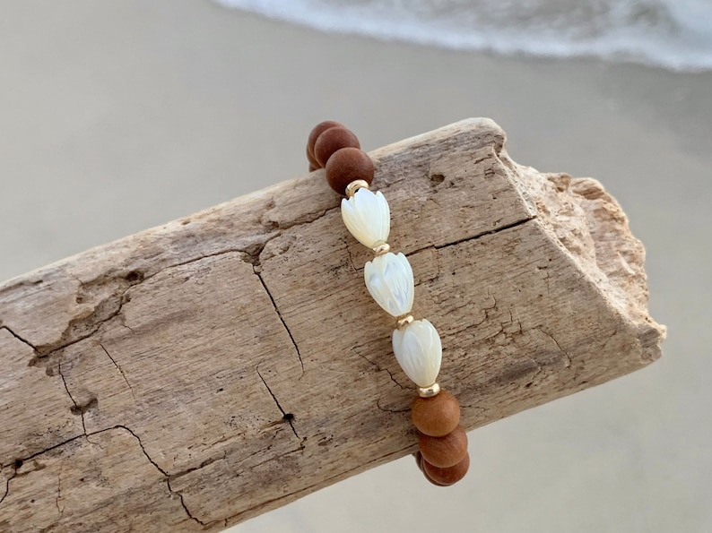 Peut inclure: Un bracelet en bois brun perl&eacute; avec trois perles de fleurs en coquille blanche. Le bracelet est sur un morceau de bois flott&eacute;.