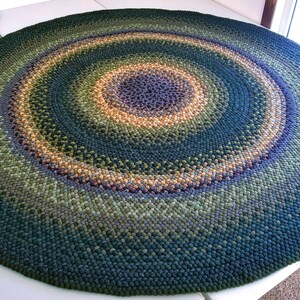 May include: A large, round braided rug with concentric circles of color. The rug transitions from a deep teal outer ring to shades of green, gold, and purple in the center. The rug is a decorative floor covering.